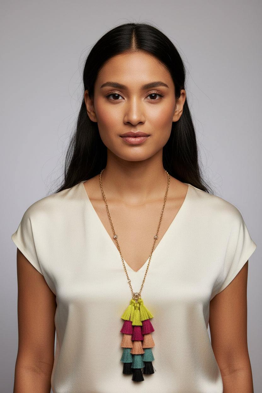 A gold chain necklace with a tiered tassel pendant in bright yellow, magenta, peach, teal, and black, displayed on a hot pink jewelry bust against a dark gray background.
