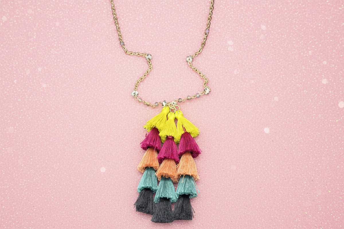 A gold-tone chain necklace with a tiered tassel pendant in bright yellow, magenta, peach, teal, and black, displayed against a dark gray background with a pink jewelry stand partially visible at the top.